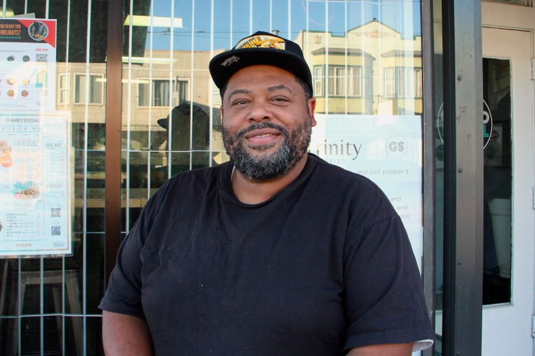 A man wearing a black t-shirt and cap sits and smiles outside a Bayview storefront with a menu and signs visible behind him.