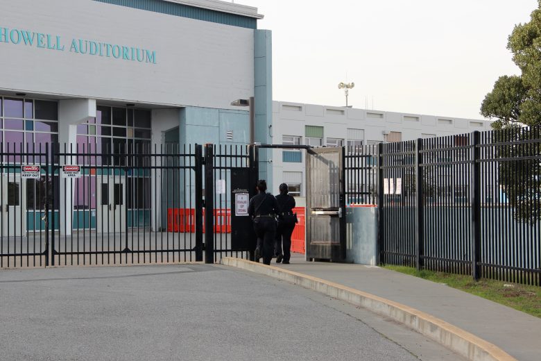 Three people in dark uniforms walk through a gate next to a building labeled "Howell Auditorium.
