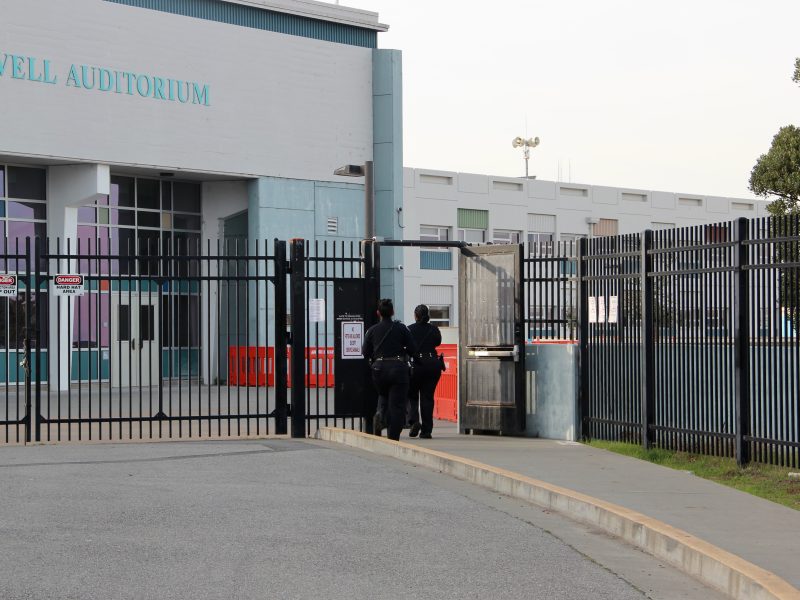 Three people in dark uniforms walk through a gate next to a building labeled "Howell Auditorium.