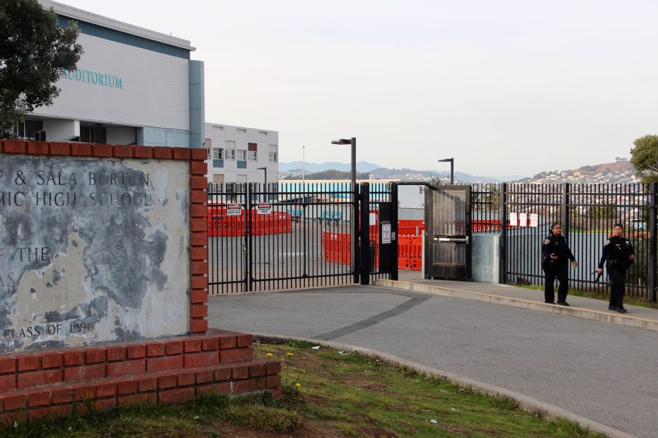 Two security officers stand near the entrance gate of a high school campus, with a faded sign and school buildings visible in the background.