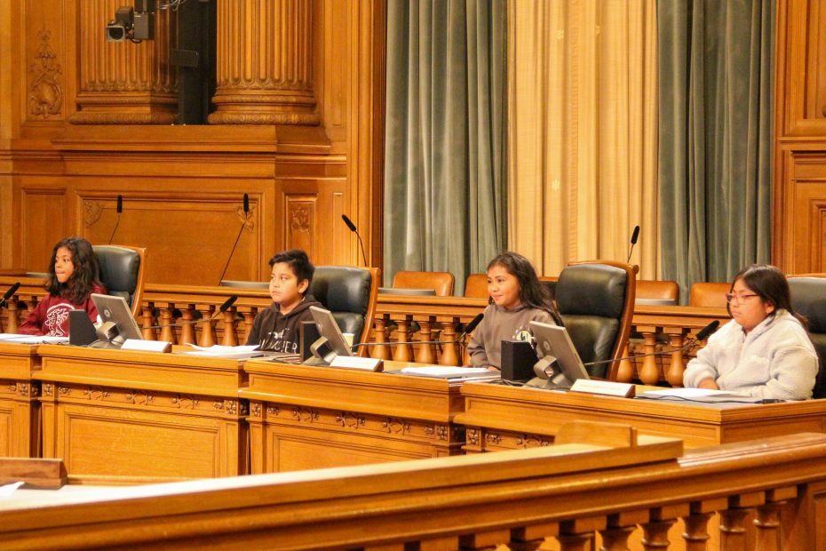 Four young people sit at a panel desk with microphones and monitors in a wood-paneled room, possibly a government or council chamber.