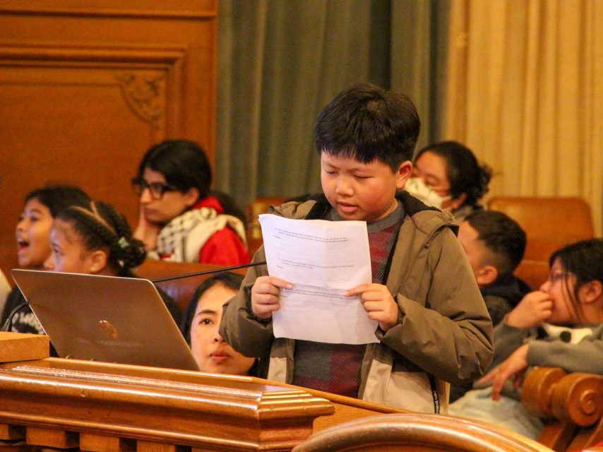 A young boy stands at a podium reading from a sheet of paper in a room with several seated children in the background.