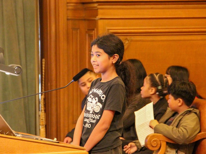 A young student stands and speaks at a podium with a microphone in a wood-paneled room, while other children sit and listen in the background.