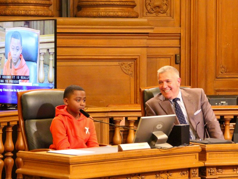 A boy in an orange hoodie speaks into a microphone at a wooden desk, while a man in a suit sits beside him smiling; a screen displays the boy’s image.
