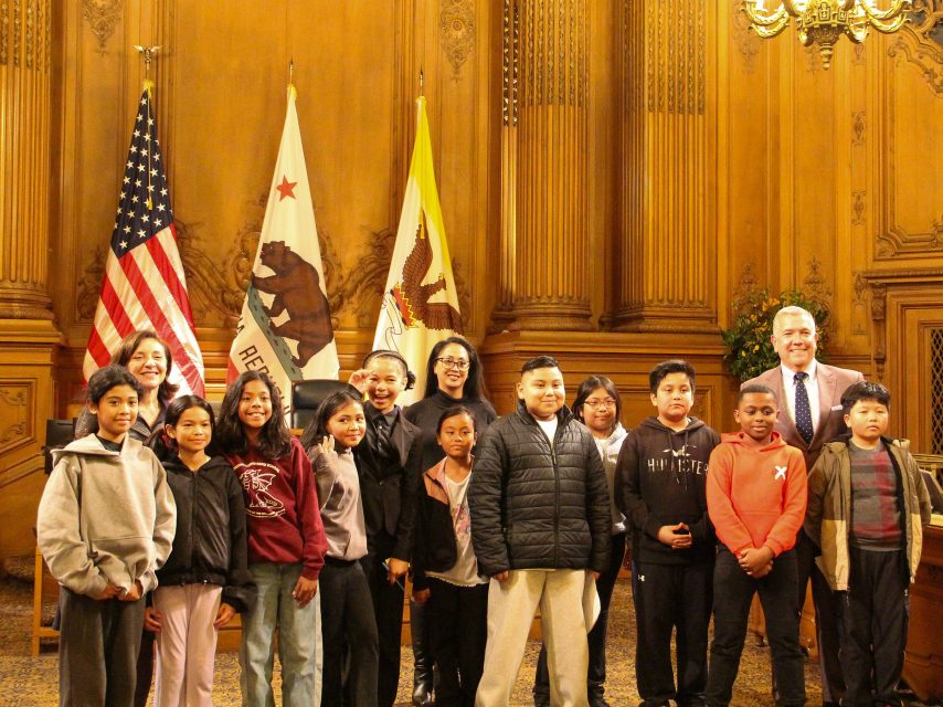 A group of children and four adults pose for a photo inside a wood-paneled room with U.S. and California flags in the background.