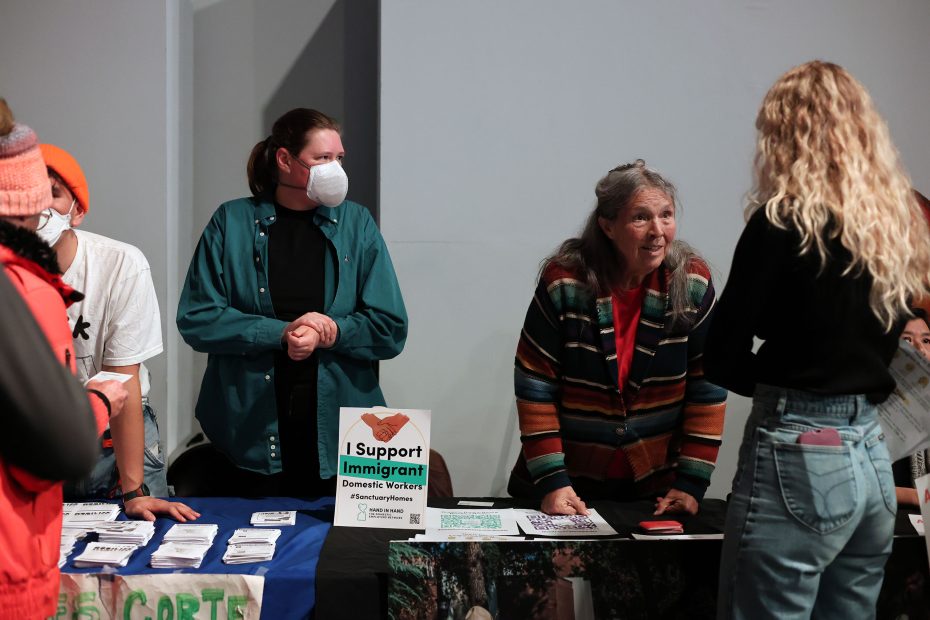 Several people stand and talk at a table displaying pamphlets and a sign reading "I Support Immigrant Domestic Workers.