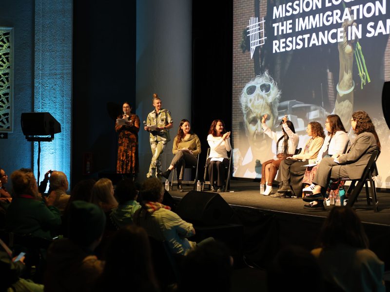 A panel of seven people sit on stage discussing immigration at an event, with an audience seated in front of them and a large screen displaying the topic behind them.