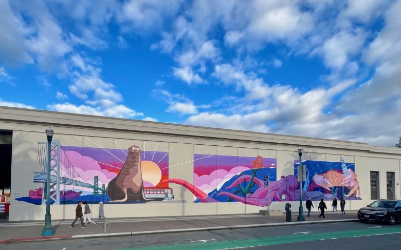 A mural on a building wall depicts a sea lion, a bridge, a cable car, and abstract waves under a blue sky with clouds. People walk on the sidewalk below.