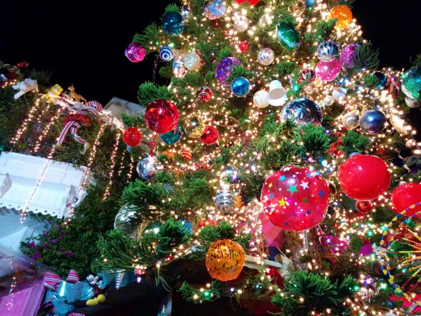 A decorated Christmas tree with colorful ornaments and lights is displayed outdoors at night, with festive lights and decorations on nearby buildings.