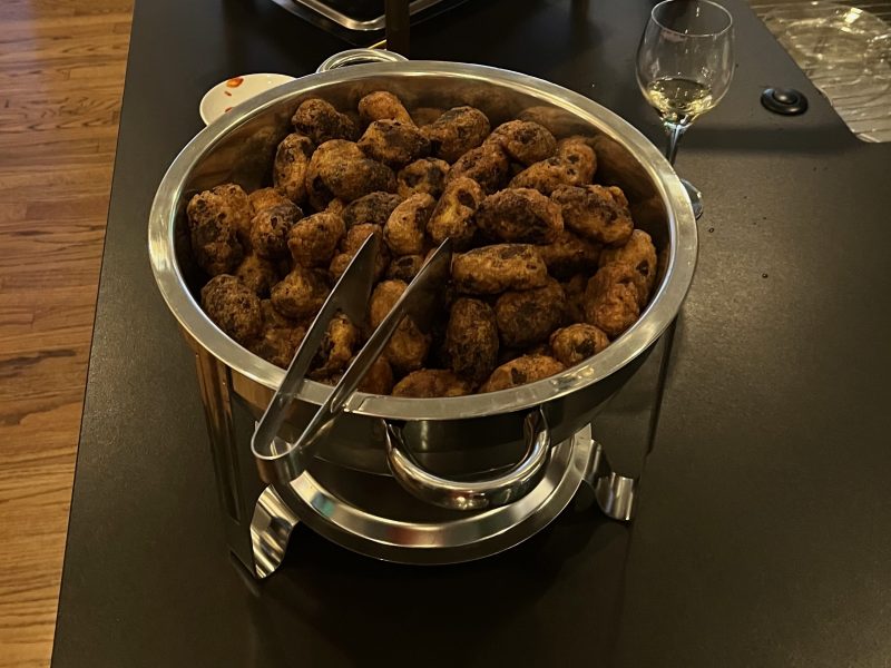 A stainless steel chafing dish filled with fried food items, possibly croquettes or fritters, with serving tongs and a wine glass nearby on a dark countertop.