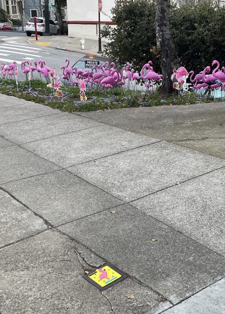 A group of pink plastic flamingo lawn ornaments is displayed on a patch of grass near a sidewalk, with some cartoon cutouts and a yellow sidewalk tile featuring a pink flamingo.