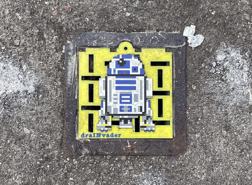 A pixel art depiction of R2-D2 on a yellow background is fixed to a metal grate on a concrete surface with the text "drallnvader" at the bottom.