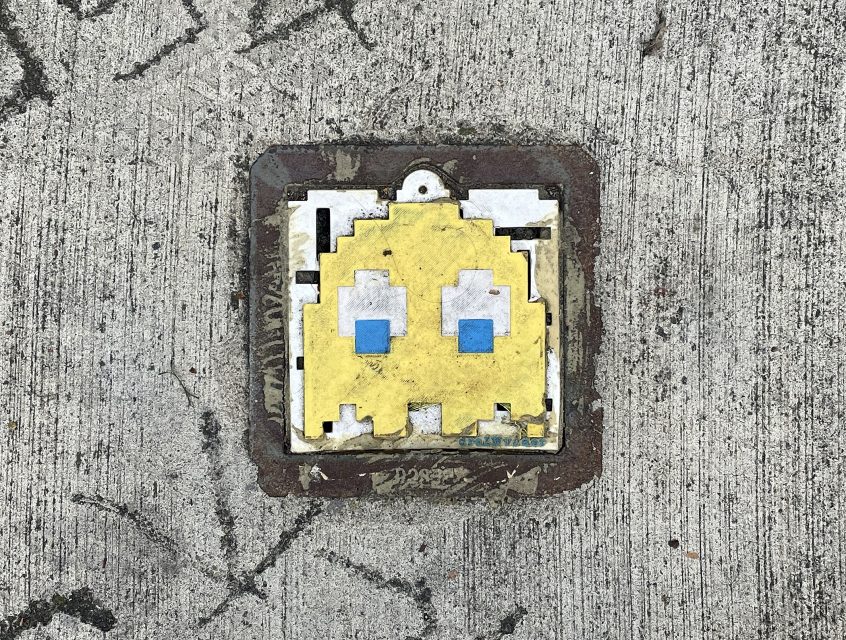 A tile embedded in concrete depicts a pixelated yellow ghost with blue eyes, resembling a character from the Pac-Man video game.