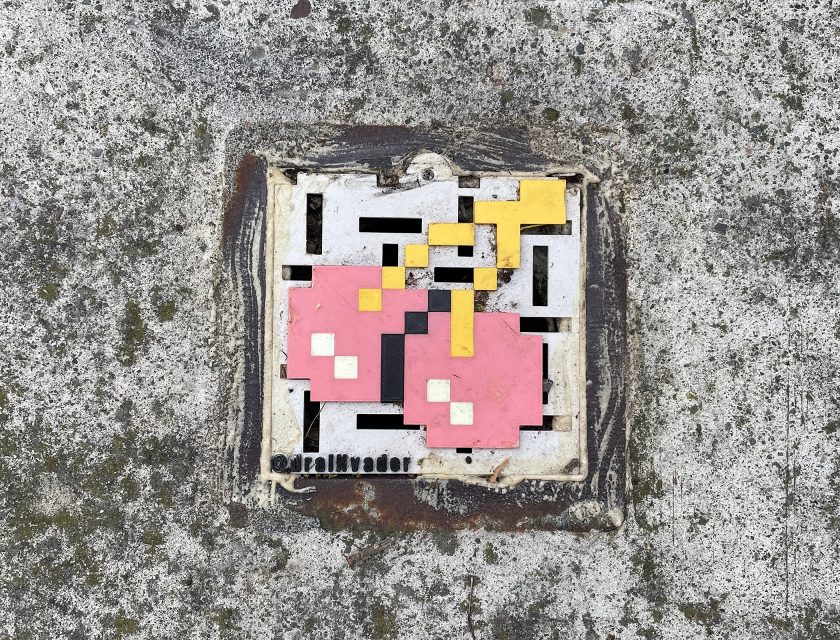 Mosaic street art of two pixelated pink cherries with yellow stems on a concrete surface, tagged with "@realinvader.