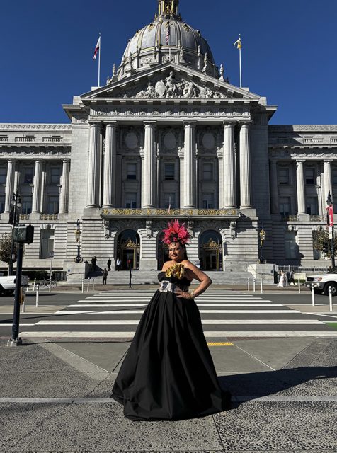 A person in a black gown and elaborate red headdress stands in front of a grand building with columns and a dome on a sunny day.