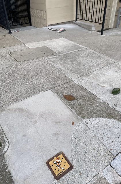 A small tile artwork is embedded in the sidewalk, with a few leaves and scattered debris visible on the surrounding pavement.
