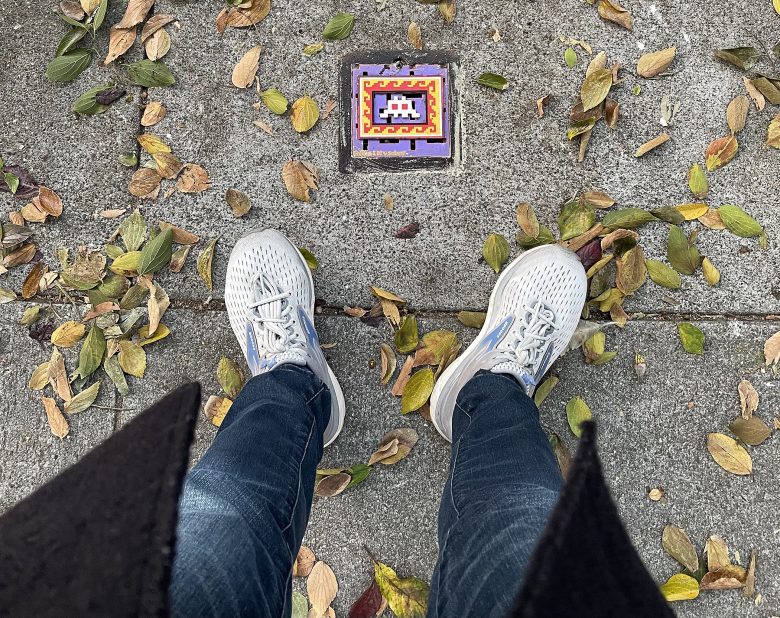 Person standing on a leaf-strewn sidewalk, wearing white sneakers and blue jeans, facing a colorful mosaic tile with a pixel art design embedded in the pavement.
