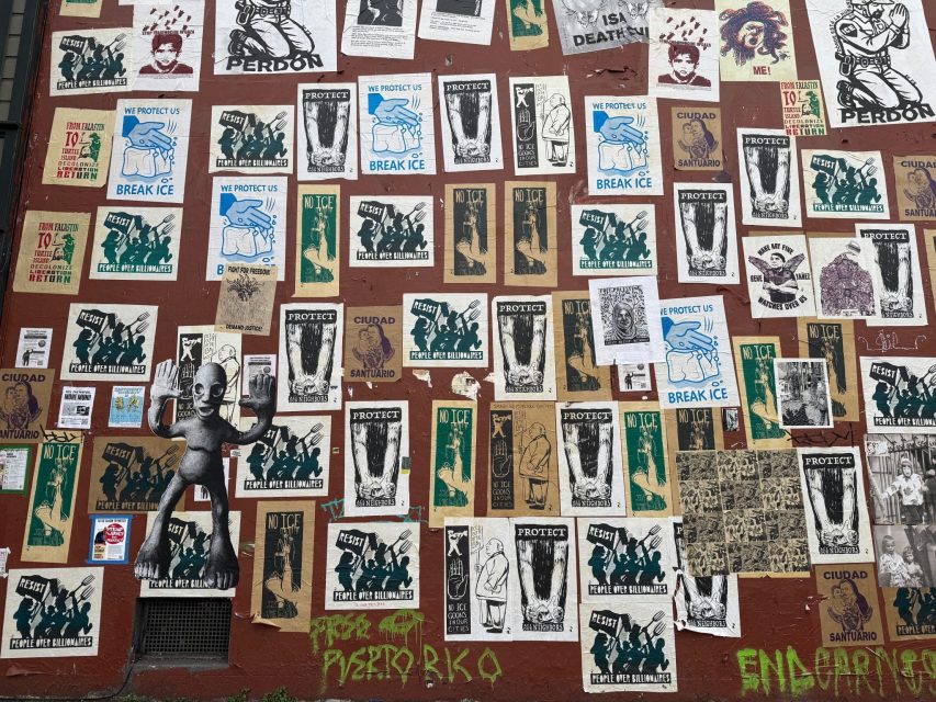 A red wall covered with various posters, including “PROTECT US,” “BREAK ICE,” and several black-and-white illustrations. At bottom left, there is a small metallic alien figure.