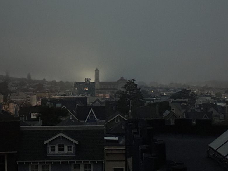 Rooftops of houses at dusk with a foggy sky, distant tower illuminated by a light, and low visibility over the city.