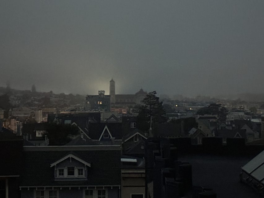 Rooftops of houses at dusk with a foggy sky, distant tower illuminated by a light, and low visibility over the city.