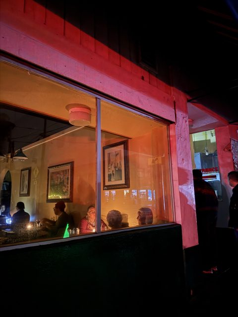 People sit inside a restaurant at night, seen through a window with colorful red and orange lighting reflecting on the walls.