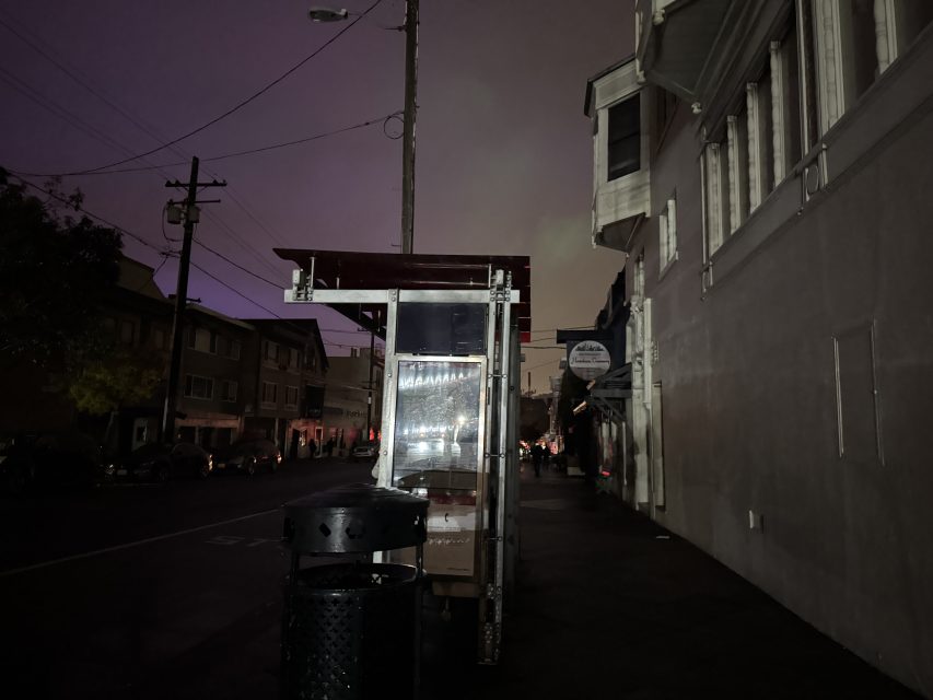 A dimly lit bus stop shelter on a dark street at night, with surrounding buildings and power lines visible under a cloudy sky.