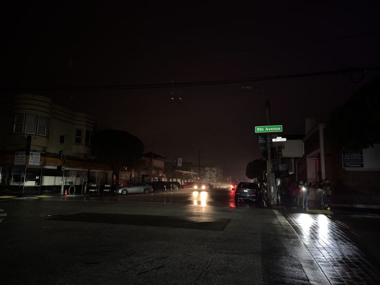 A dark, wet street at night with cars and headlights in the distance; a group of people stands under a glowing 9th Avenue street sign.