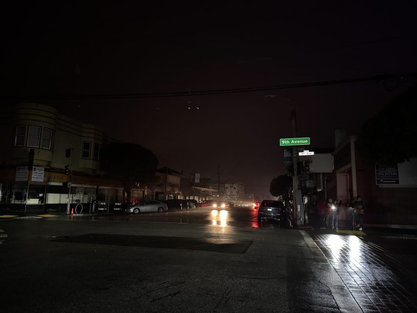A dark, wet street at night with cars and headlights in the distance; a group of people stands under a glowing 9th Avenue street sign.
