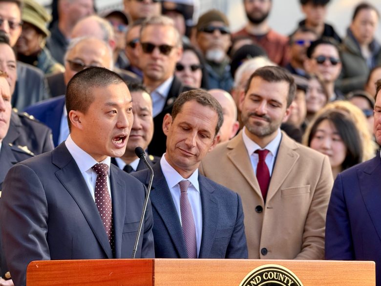 A man in a suit speaks at a podium with a seal, while a crowd of people, some in suits and coats, listen behind him outdoors.