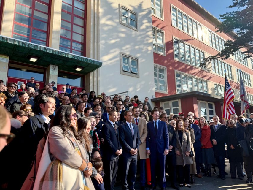 A large group of people, including officials in suits, pose for a photo outside a multi-story school building with flags visible.