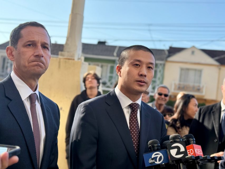 Two men in suits stand in front of microphones from news outlets, speaking outdoors with a group of people and houses in the background.
