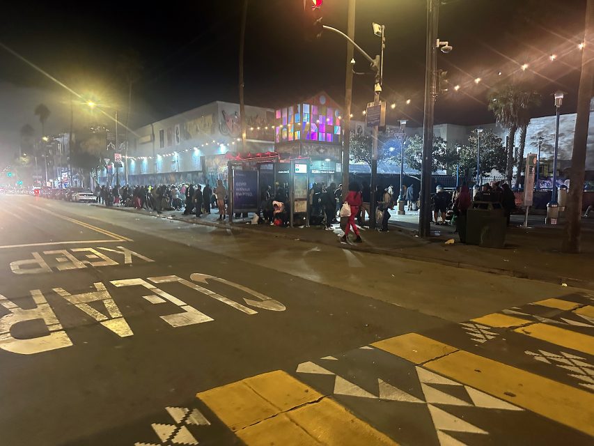 A crowd of people lines up outside a building at night near a brightly lit bus stop; street markings and palm trees are visible in the scene.