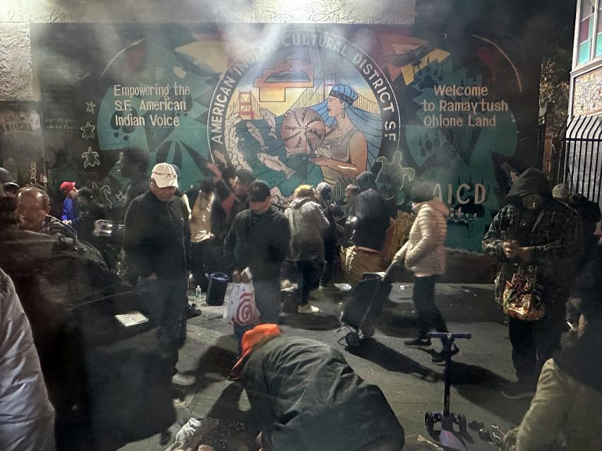 A group of people gather at night in front of a mural in San Francisco’s American Indian Cultural District; some appear to be receiving goods or services.