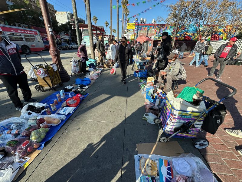 Street vending thrives as people sell food and goods laid out on blankets and carts along a busy sidewalk in an urban area, with pedestrians walking by.