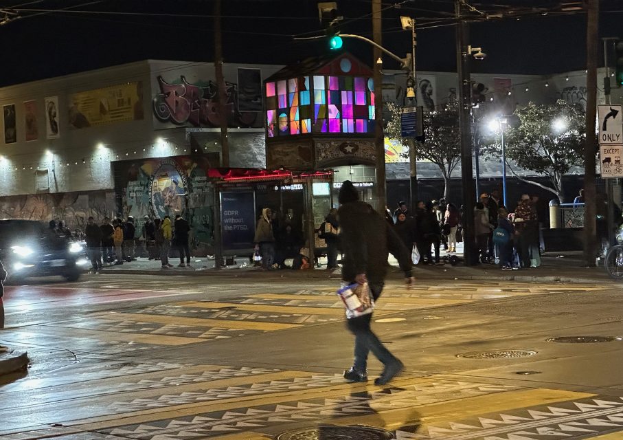 A person walks across a city street at night carrying a box, with a crowd and colorful illuminated building in the background.