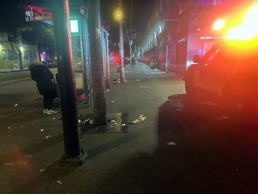 A city street at night with scattered trash, a police car with flashing lights, and a person standing near a bus stop.