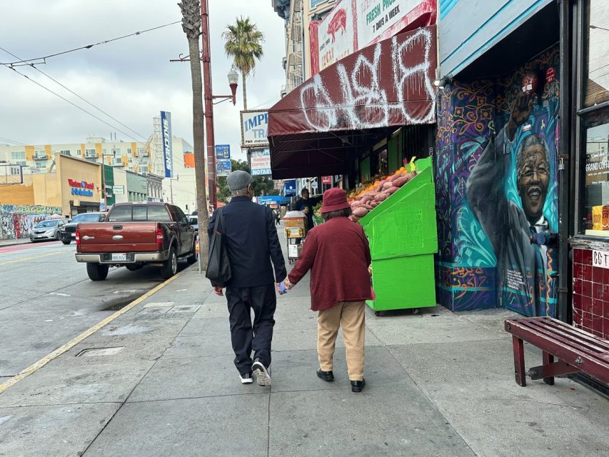 Two people walk hand-in-hand on a city sidewalk past a produce stand and a mural featuring a smiling man. The street is lined with shops, parked cars, and graffiti.