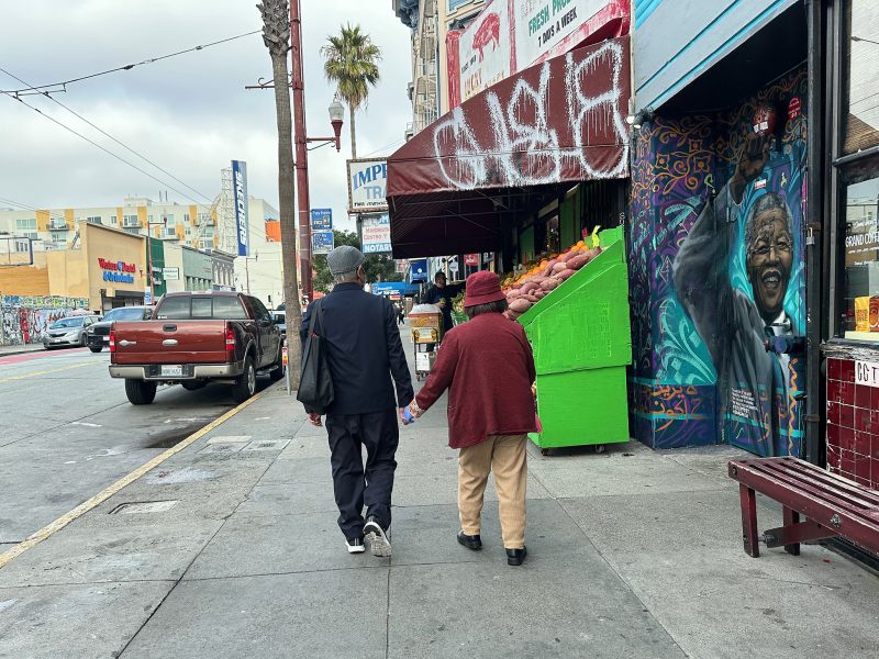 Two people walk down a city sidewalk past a produce stand with graffiti and a mural of a smiling person on the wall.