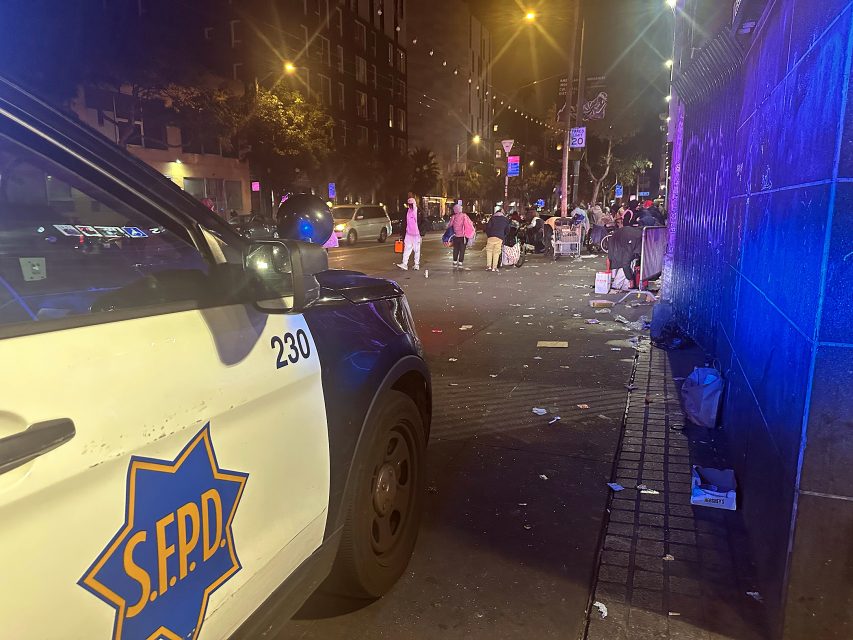 A San Francisco police car is parked on a littered city street at night as people gather near a crowded sidewalk.