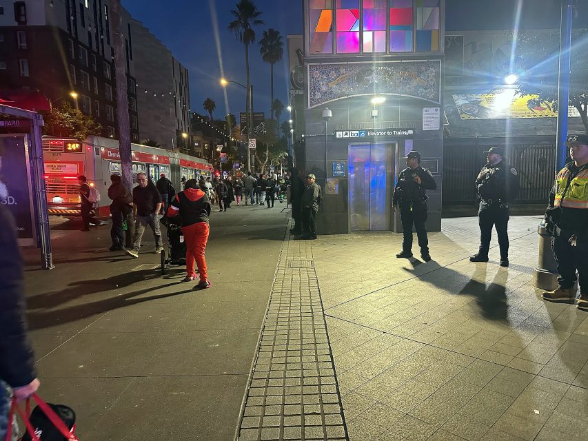 People stand and walk near emergency vehicles and uniformed officers outside a building entrance at dusk in an urban setting.