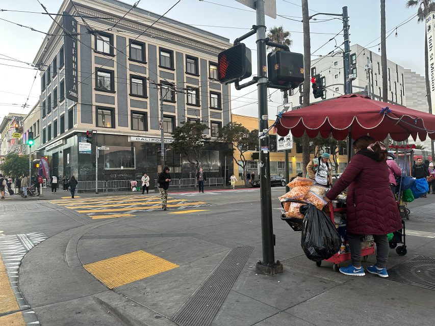 A street vendor stands by a cart of goods under a red canopy at a city intersection, with pedestrians and a large building in the background.