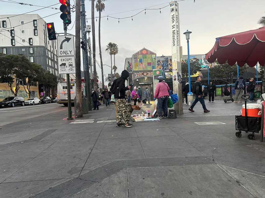 Street scene at an urban intersection with people walking, a street vendor displaying goods on the ground, traffic lights, buildings, and palm trees in the background.