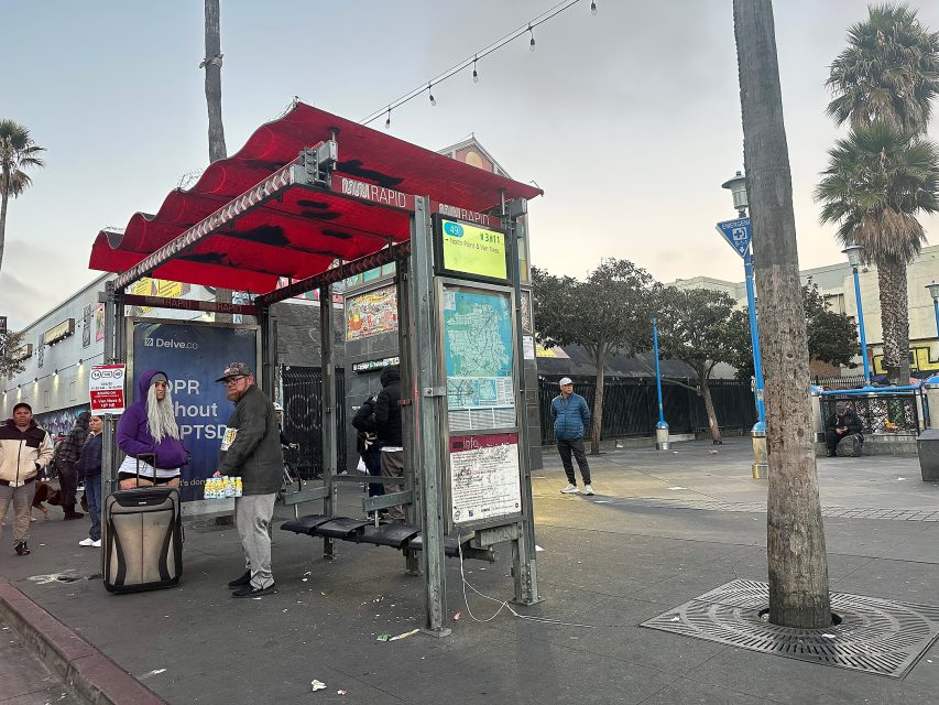 People stand at a red-roofed bus stop next to a street, with palm trees and a billboard in the background on a cloudy day.