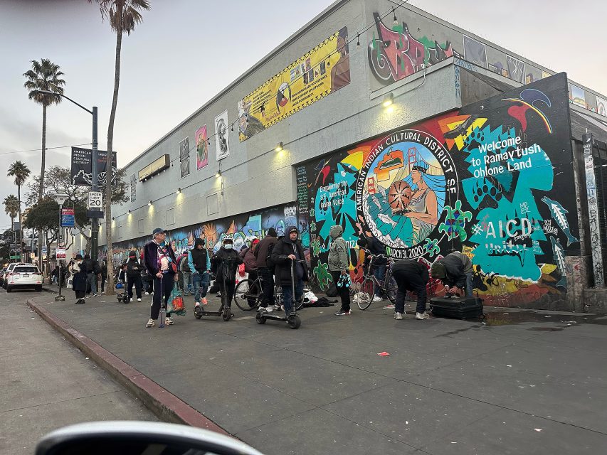 People gather on a city sidewalk in front of a building with colorful murals and graffiti art, including a sign reading "Welcome to Xicanx/Latinx Cultural District.