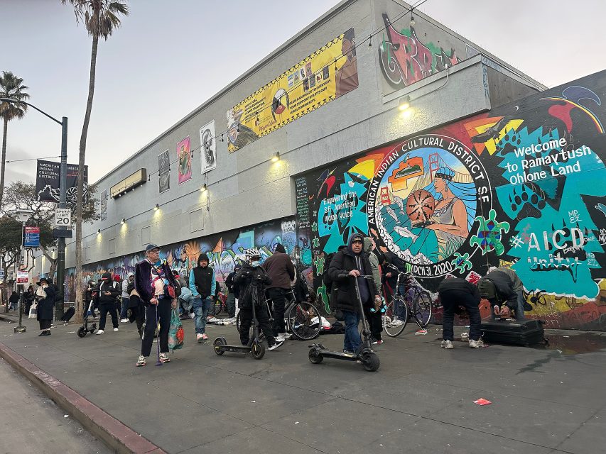 People gather on a city sidewalk with bikes and scooters in front of a mural and graffiti-covered building. A street sign, palm tree, and banners are visible.