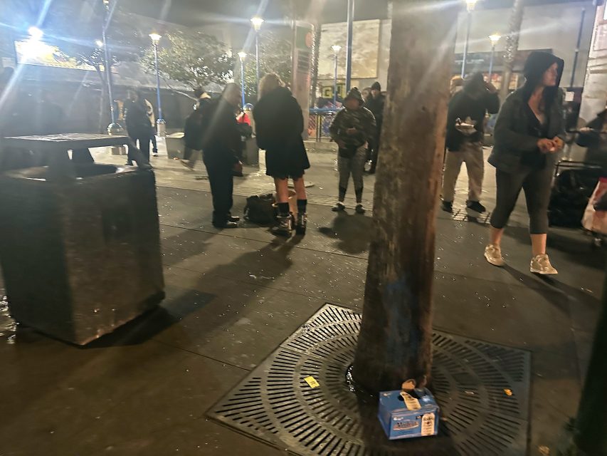 A group of people stand on a dimly lit city sidewalk at night near a tree and a trash bin, with scattered items on the ground.