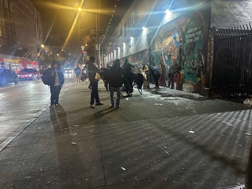 People stand and walk along a city sidewalk at night near a mural-lit wall, with parked cars and a bus visible in the street.