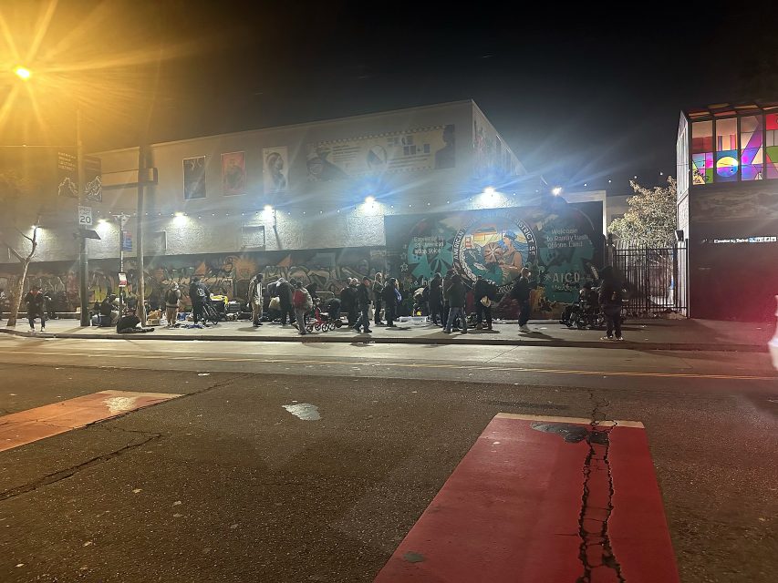 A large group of people gathers on a sidewalk at night in front of a mural-covered building, illuminated by streetlights and passing car headlights.