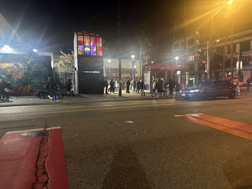 A nighttime street scene shows people gathered near a brightly lit bus stop with a mural and colorful cube structure in the background as cars pass by.