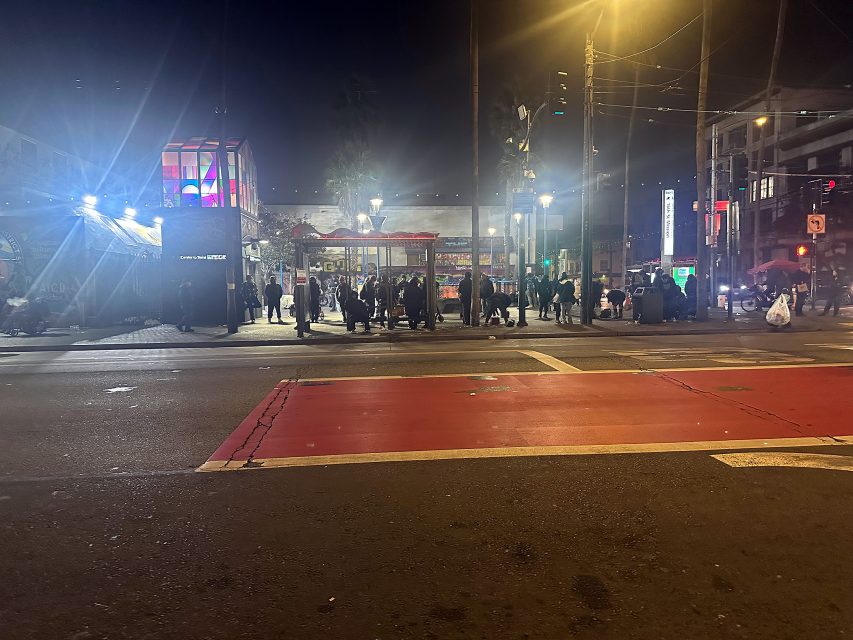 A nighttime street scene shows a group of people gathered on the sidewalk near illuminated buildings and streetlights, with cars and a red-painted crosswalk in the foreground.
